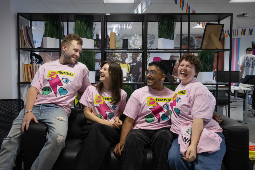 Four members of the Manchester Pride team wearing pink t-shirts with their colourful logo on the front, smiling and laughing on a sofa.